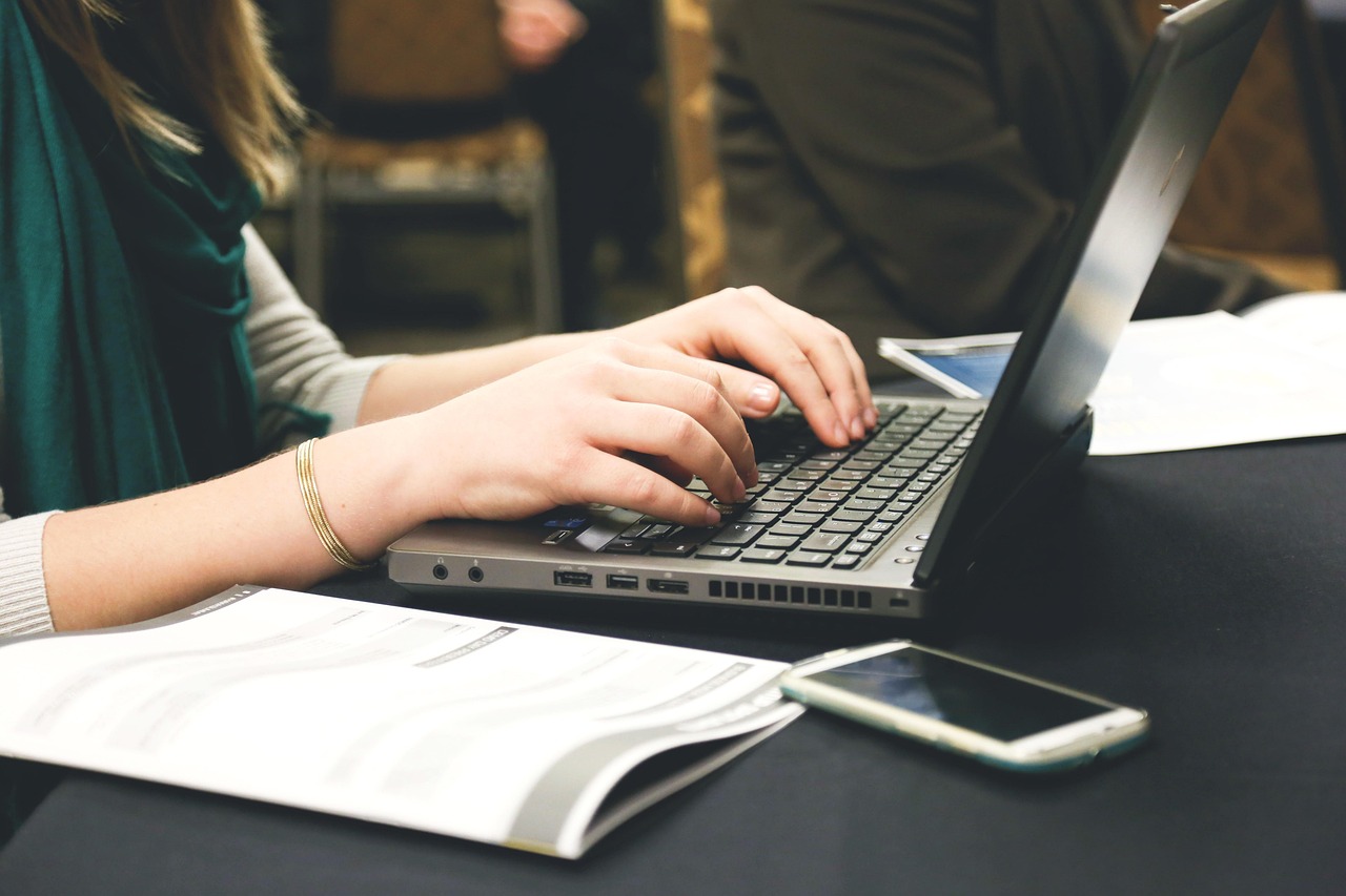 Mains qui tapent sur le clavier d'un ordinateur portable, à côté d'un carnet ouvert et d'un téléphone
