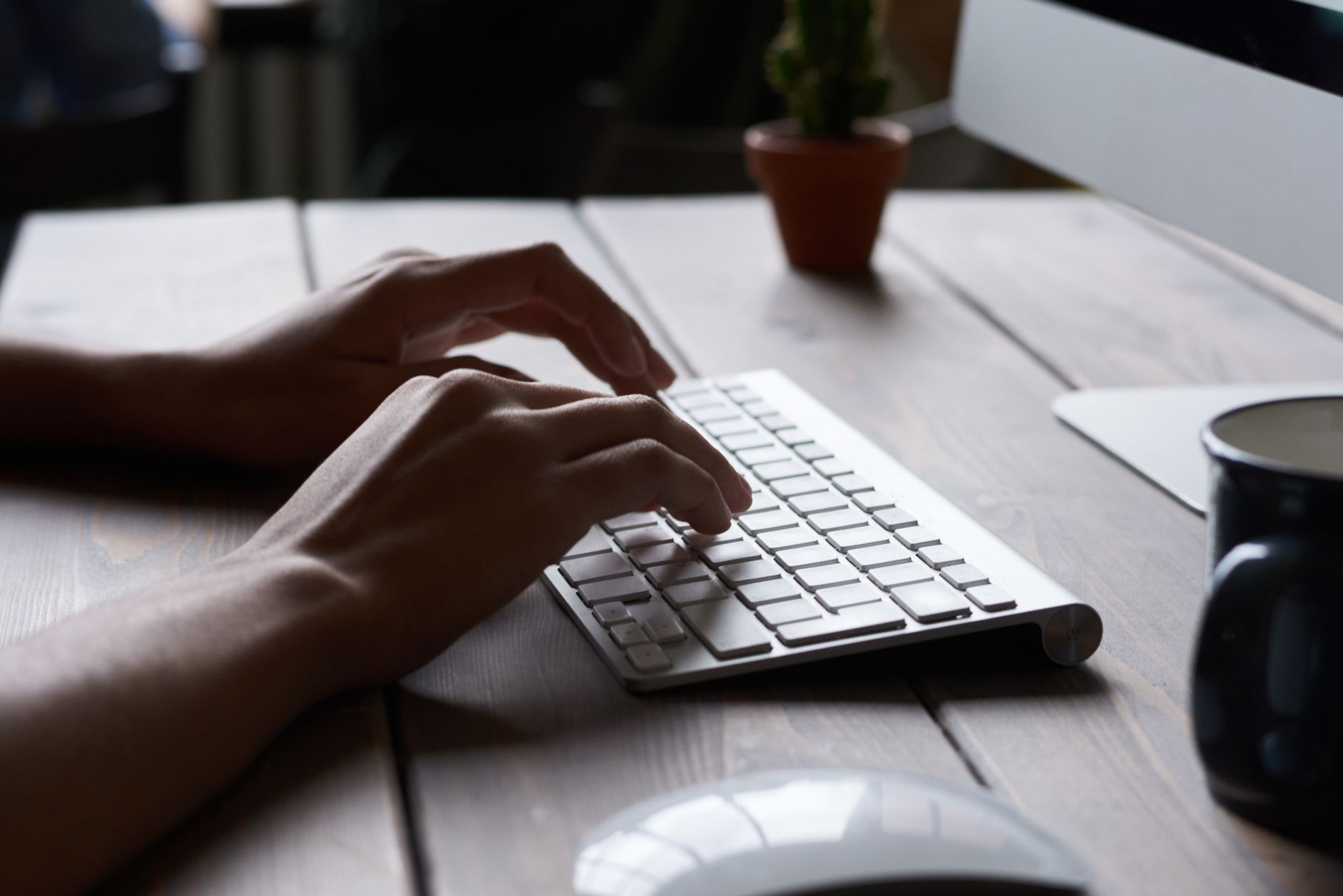 Mains tapant sur un clavier sans fil blanc posé sur un bureau en bois, avec une souris, une tasse et un petit cactus.