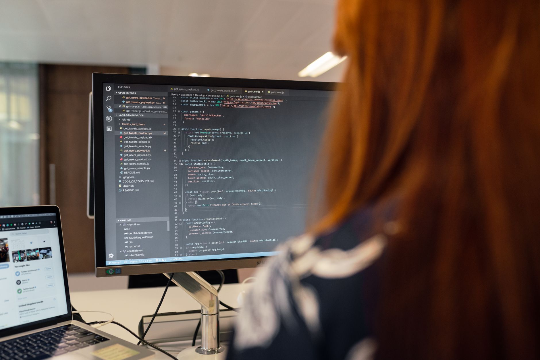 Personne vue de dos devant un écran affichant du code dans un éditeur, avec un ordinateur portable à côté dans un bureau.